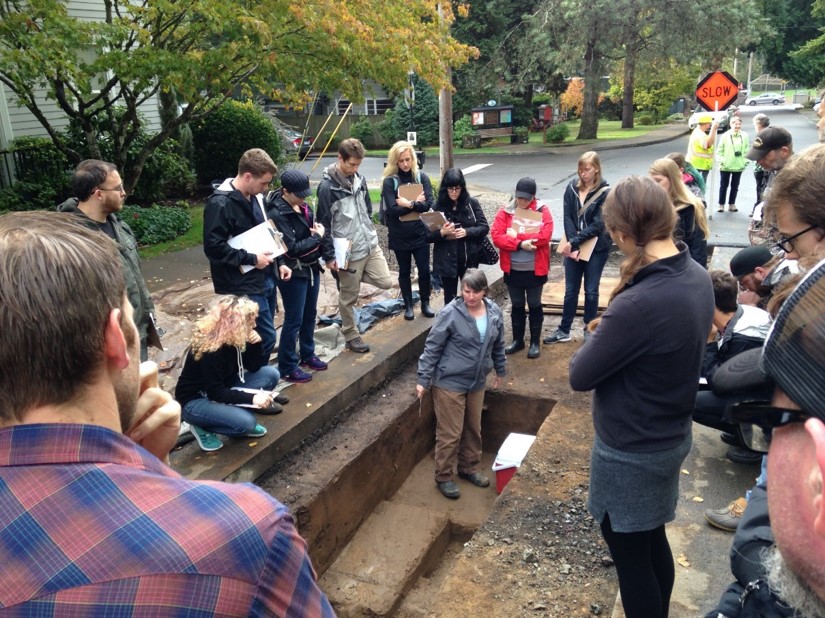 Students gathered around an excavation unit in the middle of a neighborhood street