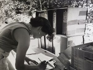 Black and white photo of an archaeologist doing paper work at a field desk stacked with binders and folders.