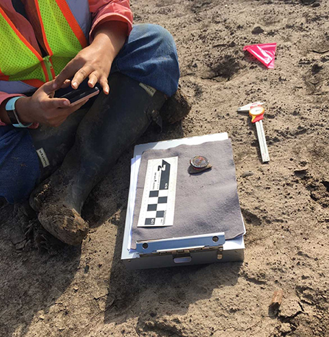 An archaeologist sits cross-legged on the ground with a clipboard and scale in front of them, measuring artifacts