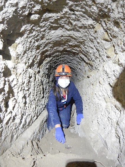 a person in a hard hat and mask crawling through a rocky tunnel, photo copyright Cocceius Association